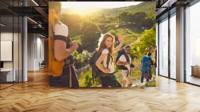 Group of happy backpackers trekking on sunny day. Young tourists traveling and enjoying active summer vacation. Smiling woman looking at camera and waving hand walking down hiking trail with friends Wall mural