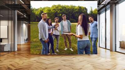 Group of happy friends or corporate coworkers standing on grass in summer park on weekend and listening to manager with clipboard listing their tasks and explaining rules of outdoor team building game Wall mural