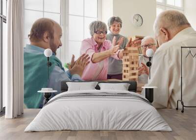 Group of happy senior people having fun and playing board games in retirement home. Several cheerful old men and women gather around table and take turns to take wood blocks out of wooden tower Wall mural