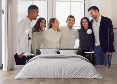 Group of happy smiling young diverse people sitting on windowsill or standing by window and hugging. Friends, college or university students, coworkers, or business startup team gathered all together Wall mural