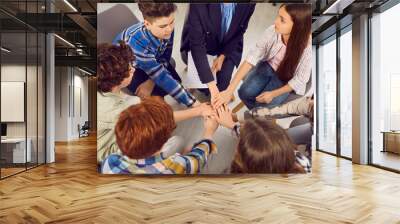 Group of school children learning what teamwork and support are. Team of student boys and girls together with woman teacher sitting in circle in classroom and holding their hands together Wall mural