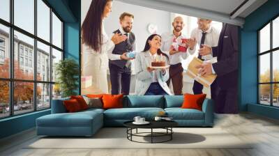 Group of young smiling business people congratulating their happy female colleague on her birthday giving her birthday cake and present gift boxes on workplace in office. Corporate party Wall mural