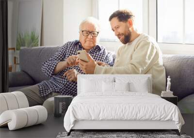 Happy adult son helps elderly father understand the phone and modern technology, sitting together on the sofa at home. Child learning old parent about the internet and online tools. Wall mural