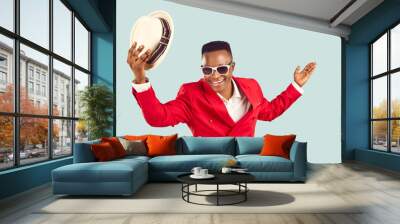 Happy African man in funky outfit welcomes you at party. Cheerful Tanzanian gentleman in stylish red suit standing isolated on blue background takes off his hat, smiles and makes bow. Fashion concept Wall mural