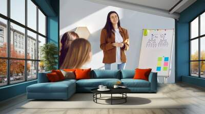 Happy beautiful Caucasian woman making presentation in business meeting. Team of people sitting in modern office room interior and listening to speaker giving master class on corporate management Wall mural