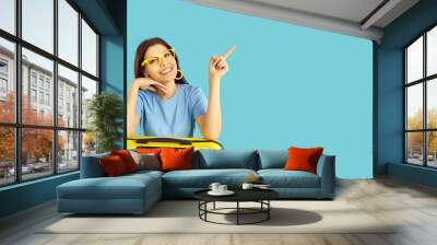 Happy beautiful young woman in glasses leaning on suitcase, smiling and pointing index finger at blank empty copyspace on right side on blue studio background. Traveling agency, holiday trip concept Wall mural