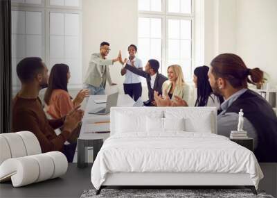 Happy business team celebrating good work results. Young ethnic man gives high five to colleague while diverse mixed race teammates sitting at office table are applauding. Teamwork and success concept Wall mural