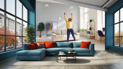 Happy cheerful young African American woman in casual clothes enjoying free time at home, standing in living room with sofa, raising hands up, stretching, smiling and charging with positive energy Wall mural