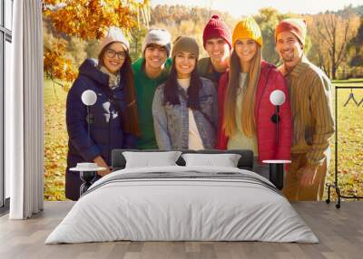 Happy cheerful young friends enjoying fall season together. Group portrait of joyful young people in colorful warm jackets, coats and hats standing in sunny autumn park, looking at camera and smiling Wall mural