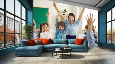 Happy children and educator raising hands up sitting at table in modern classroom. Website header with group portrait of cheerful school teacher and first grade students having fun activities in class Wall mural