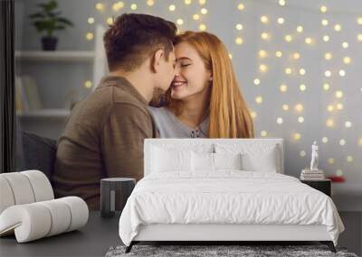 Happy couple in love sitting on sofa at home, holding gift box, smiling and touching noses. Young woman kissing boyfriend and thanking him for Saint Valentine's Day or relationship anniversary present Wall mural