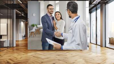 Happy couple meeting family doctor medical specialist in office. Clinician handshaking male patient hand at meeting. Male doctor talking to young husband and wife at health center or hospital Wall mural