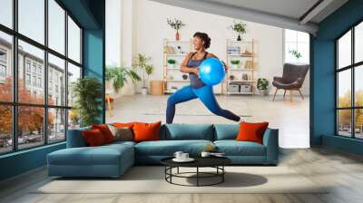 Happy energetic African American woman enjoying active workout at home. Cheerful joyful smiling young attractive female athlete doing sports exercises with fitness ball in living room interior Wall mural