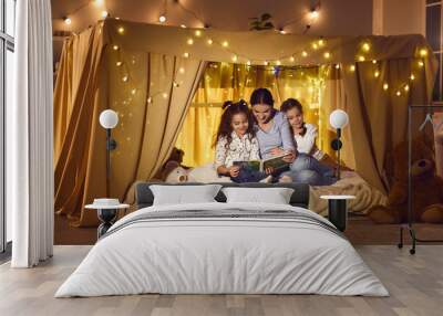 Happy family enjoying a good quiet evening at home. Mommy telling fairy tales to children. Young mother and two little daughters reading book of bedtime stories sitting in cozy playroom tent together Wall mural