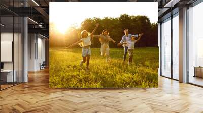 Happy family running and having fun in summer park enjoying sunny day in nature. Mother, father and their cheerful children boy and girl walking on green grass outdoors at sunset together. Wall mural