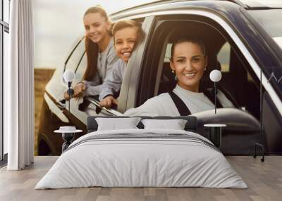 Happy family traveling by automobile. Single mother and her children enjoying road trip together. Beautiful woman driver and kids sitting inside good modern black car, looking out window and smiling Wall mural