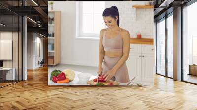 Happy fit young woman cooking healthy vegetarian food at home. Smiling housewife or diet coach standing at kitchen table and chopping red sweet or bell peppers for vegetable salad rich in vitamins Wall mural