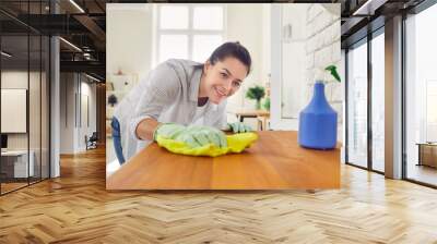 Happy housewife cleaning up at home. Young woman in rubber gloves holding a dry rag, smiling and wiping the wooden surface of her kitchen table. Housework, domestic chores, maintaining hygiene concept Wall mural