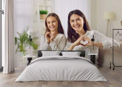 Happy loving mom and child celebrating Mother's Day together. Portrait of happy Caucasian mother and daughter sitting on sofa, looking at camera, smiling and doing heart shape gesture with hands Wall mural