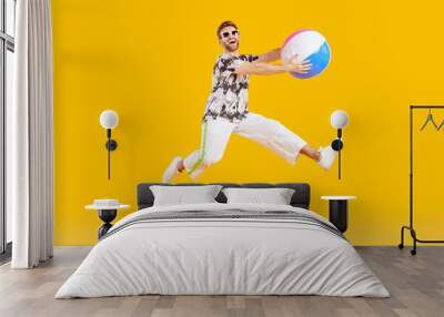 Happy man having fun during his summer holiday. Joyful guy playing with a ball at the beach. Cheerful, funny man holding an inflatable ball and jumping isolated on a yellow orange color background Wall mural