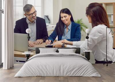 Happy married couple who are clients of bank signs paper document confirming loan for purchase of home. Female bank employee sits at desk in office and shows place for signing contract. Wall mural