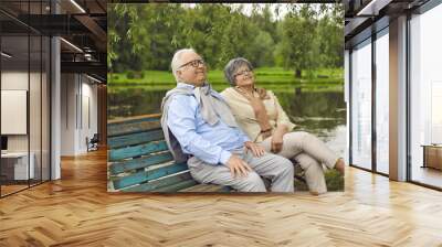 Happy married senior couple enjoying good time together. Relaxed mature man and woman sitting on an old wooden bench under a willow tree on a quiet bank of a lake or river Wall mural