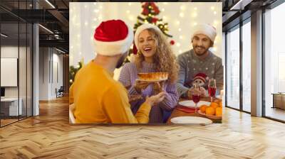 Happy millennial friends having fun and enjoying dinner at cozy Christmas party at home. Beautiful woman holding tasty cake she baked herself, smiling and giving it to man sitting at table next to her Wall mural