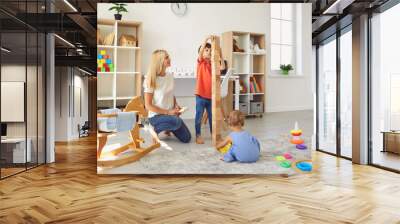 Happy mother with her two children playing with lots of toys and building block tower at home Wall mural