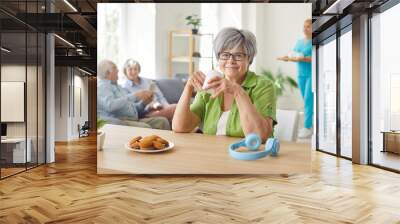 Happy old woman enjoys daily life in retirement home. Portrait of smiling gray haired lady in glasses sitting at table drinking tea, eating cookies, looking at camera, with other seniors in background Wall mural