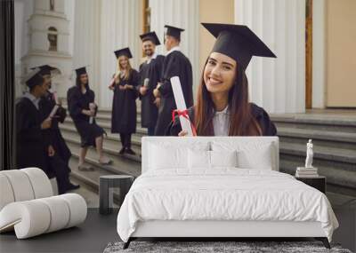 Happy positive pretty girl university graduate standing holding diploma in hand over group of mates and university building at background. Graduation from university, education, diploma concept Wall mural