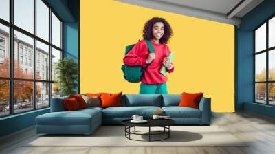 Happy school child. Cheerful kid with books and backpack. African American student girl in modern red sweatshirt and green jeans standing isolated on yellow background, looking at camera and smiling Wall mural