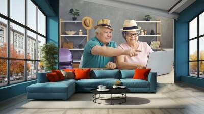 Happy senior couple at home using a laptop to pay for tickets and book a room online. Cheerful man and woman with hats on their heads ready to rest. Concept of summer vacation abroad. Wall mural