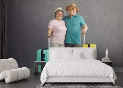 Happy senior tourists ready for vacation and adventure trip. Old retired married couple in sun hats and glasses with mint green and yellow travel suitcases standing in studio with gray background Wall mural