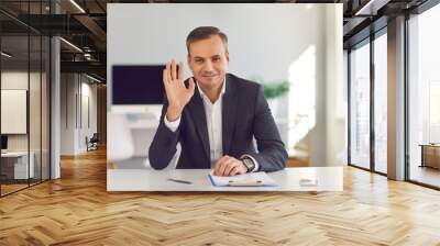 Happy smiling man in suit sitting at office desk, looking at camera and waving hand saying hello during video call. Webcam portrait of business coach, CEO, financial consultant or university professor Wall mural