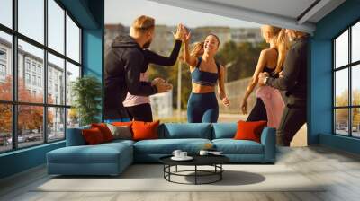 Happy smiling sporty young friends giving high five to a woman rejoicing the success in sport training after jogging exercises outdoors. People standing in the park after workout in nature. Wall mural