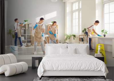 Happy smiling young cleaners men and women in uniform wiping desk, mopping floor and vacuuming. Cheerful team of professional janitors washing office workplace. Cleaning service concept. Wall mural