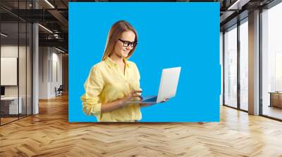 Happy student working on notebook PC. Studio shot of pretty young woman in yellow shirt and eyeglasses standing isolated on solid blue background, holding laptop computer, using touchpad and smiling Wall mural