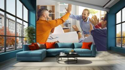 Happy successful multiracial business team clapping hands while two colleagues giving high fives gesture each other. Businesspeople laugh and cheer sitting at table in office on briefing meeting Wall mural