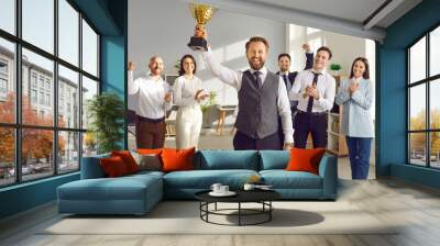 Happy winner businessman receives a business award. Joyful man holding his trophy, with a team of cheerful workers in the background celebrating and congratulating their leader on great work success Wall mural