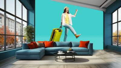 Happy woman with yellow suitcase going on summer holiday. Smiling overjoyed young girl wearing jeans, shirt and panama hat walking with her travel bag on bright turquoise background. Vacation concept Wall mural