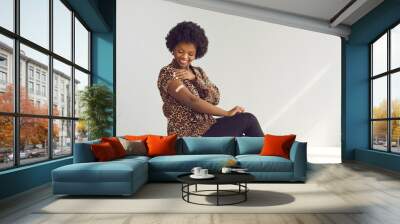 Happy young African woman shows arm after getting Covid-19 vaccine jab. Smiling woman promoting safe coronavirus (Covid19) vaccination sitting on chair on studio background with free text copyspace Wall mural