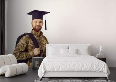 Happy young male military student wearing camouflage uniform and graduate cap standing on white copy space background, holding books and bag, looking at camera and smiling. Army and education concept Wall mural