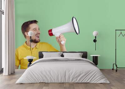 Happy young man in casual yellow T shirt standing on green colour copy space studio background, shouting through megaphone and making loud announcement about upcoming holiday sales and discounts Wall mural