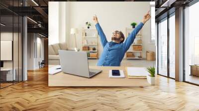 Happy young man in front of laptop computer raising hands and laughing excited about success. Emotional guy feeling glad and euphoric about getting work done and finishing successful business project Wall mural