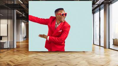 Happy young man in stylish party outfit dancing and having fun in studio. Cheerful excited African American man in red suit and sunglasses dancing isolated on light blue background. Fashion concept Wall mural