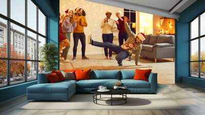 Happy young multiethnic people having fun at Christmas party in beautiful modern studio apartment. Group of excited millennial friends in Santa hats applauding funny dancing guy in ugly Xmas sweater Wall mural