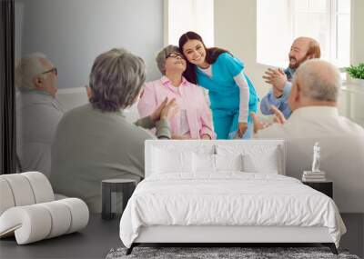 Happy young nurse and senior patients in the retirement home having fun together. Smiling girl caretaker hugs an old woman while other elderly people are clapping hands. Love, care, support concept Wall mural