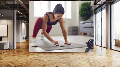 Home training. Athletic woman trains at home and puts a timer on her phone and monitors the progress of her workout on the fitness app. Concept of using modern gadgets to monitor your health. Wall mural