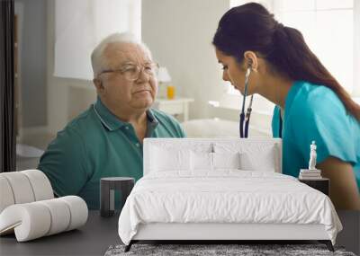 Homecare nursing service and elderly people cardiology healthcare. Close up of young hispanic female doctor nurse check mature caucasian man patient heartbeat using stethoscope during visit Wall mural