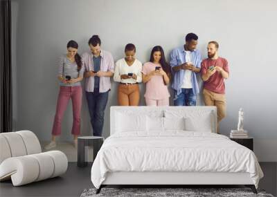 Indoor shot of diverse international group of happy young millennial people leaning on wall, looking at screens of mobile phones, connecting to free wi fi, using messengers, watching online streams Wall mural